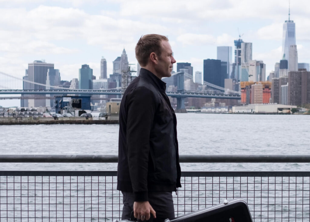 Jason stands with a suitcase by a tranquil body of water, gazing at the serene landscape before him.